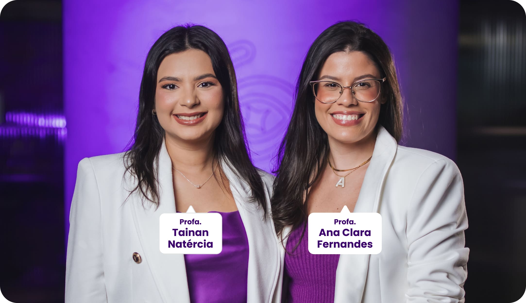Duas professoras posam sorrindo para a foto. Da esquerda para a direita: Profa. Tainan Natércia e Profa. Ana Clara Fernandes. As professoras vestem blazer branco e blusa roxa. Ao fundo, há um ambiente moderno com iluminação em roxo.