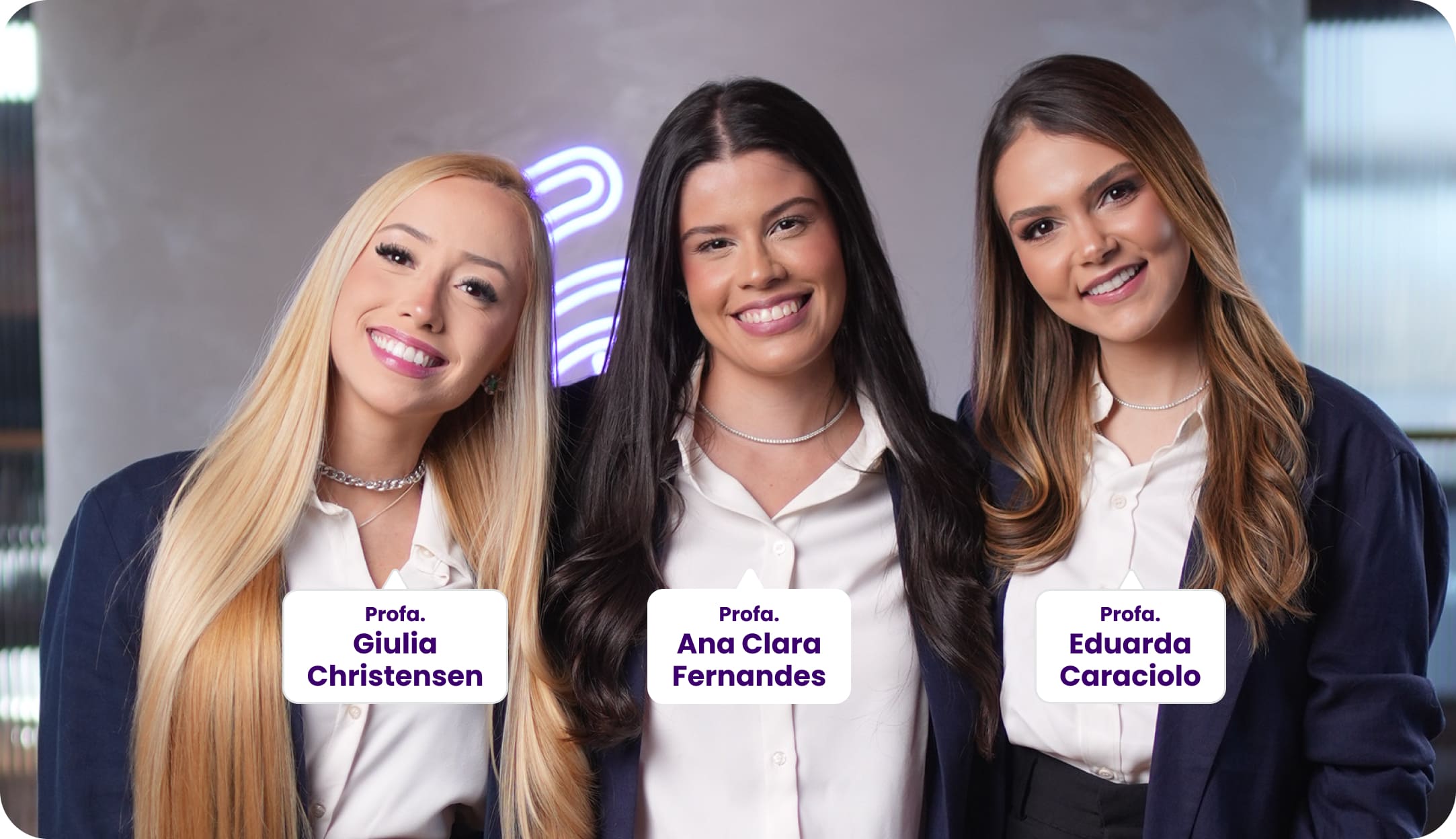 Três professoras posam sorrindo para a foto, vestindo blazers azul marinho e blusa branca. Da esquerda para a direita: Profa. Giulia Christensen, Profa. Ana Clara Fernandes e Profa. Eduarda Caraciolo. Ao fundo, um ambiente moderno com iluminação roxa difusa.