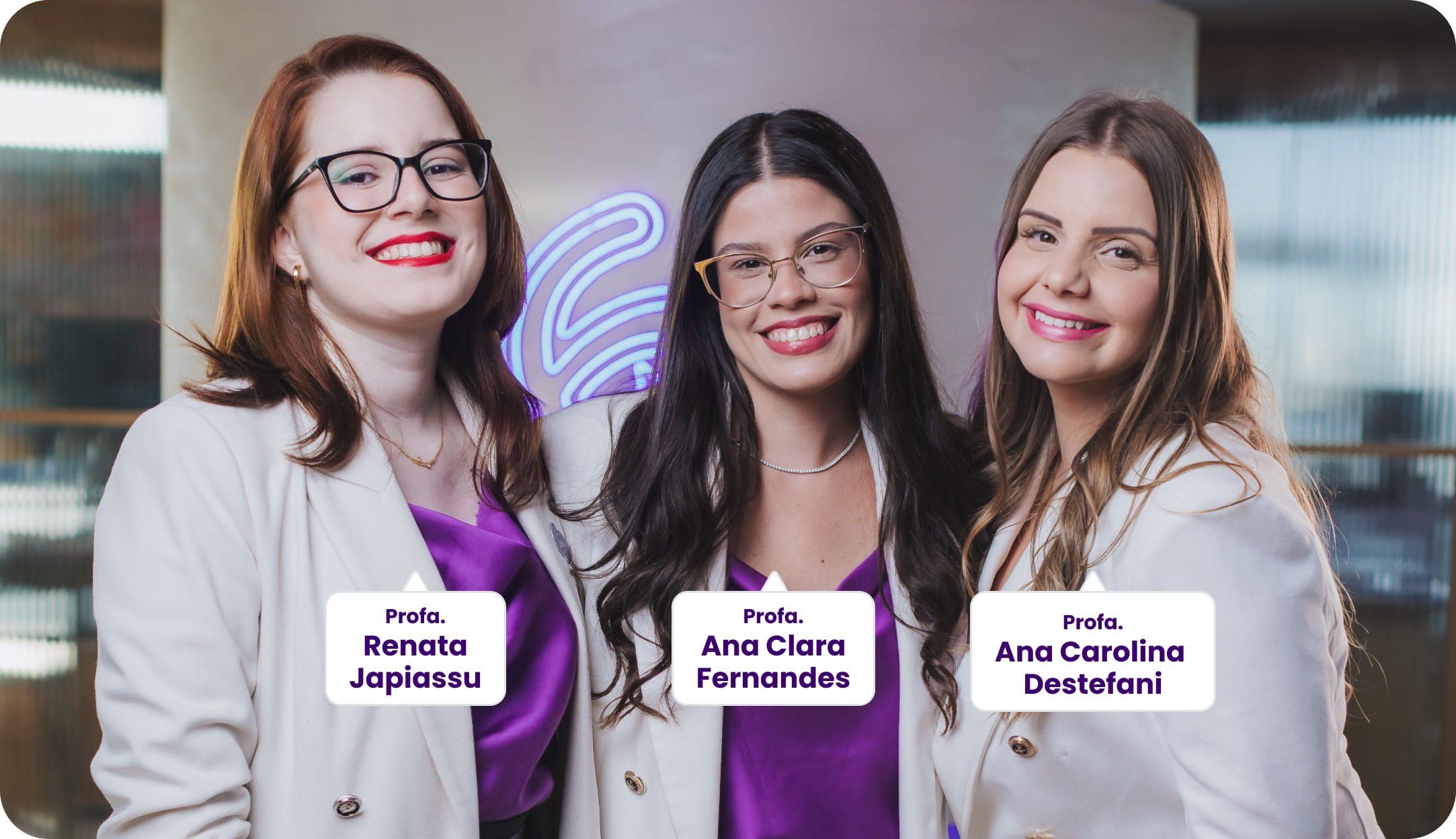 Três professoras posam sorrindo para a foto, vestindo blazers brancos e blusas roxas. Da esquerda para a direita: Profa. Renata Japiassu, Profa. Ana Clara Fernandes e Profa. Ana Carolina Destefani. Ao fundo, um ambiente moderno com iluminação roxa difusa.