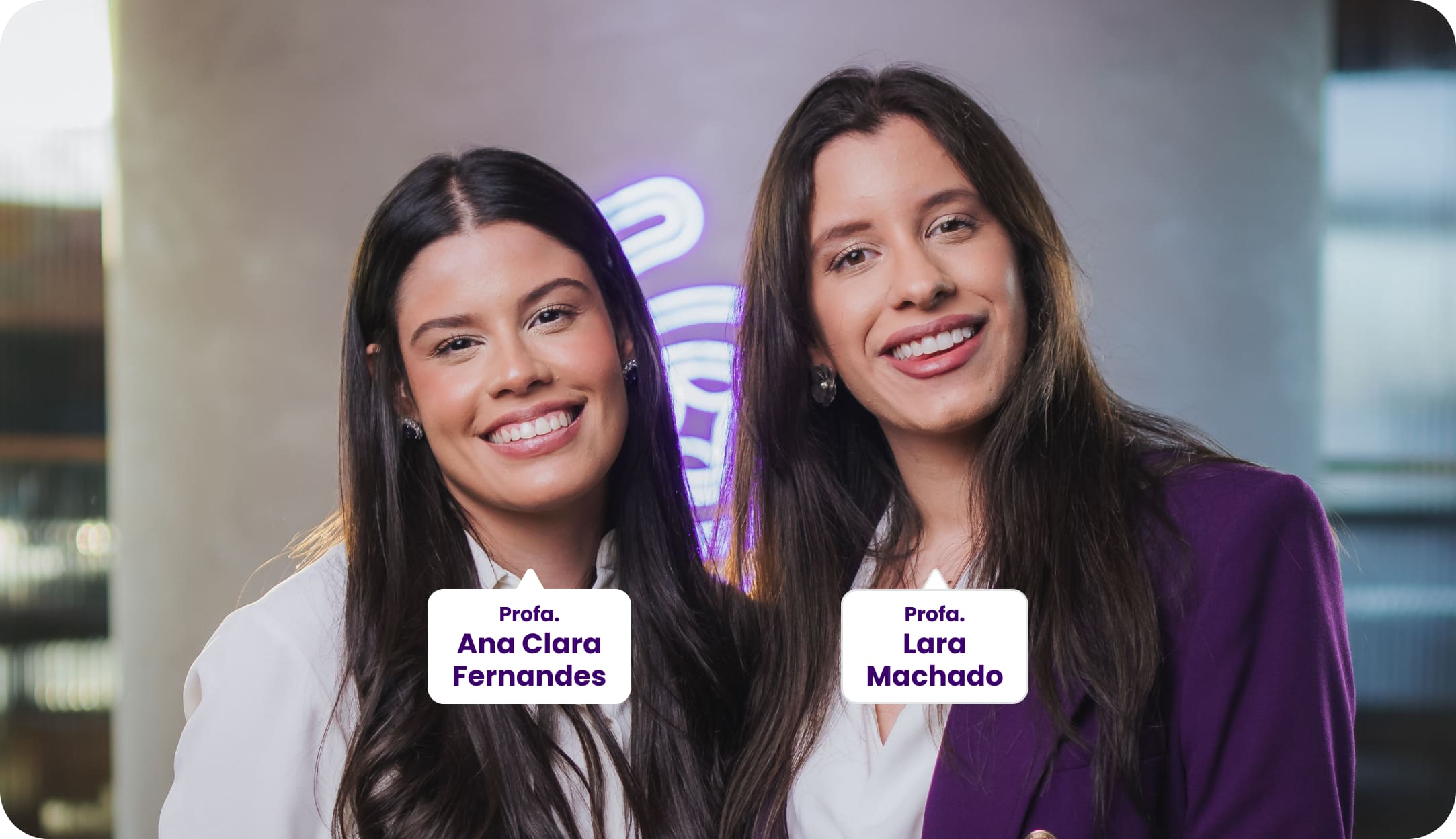 Duas professoras posam sorrindo para a foto. Da esquerda para a direita: Profa. Ana Clara Fernandes e Profa. Lara Machado. Profa. Ana Clara vestindo formalmente uma camisa branca alinhada e Profa. Lara Machado com um blazer roxo e camisa branca. Ao fundo, um ambiente moderno com iluminação roxa.